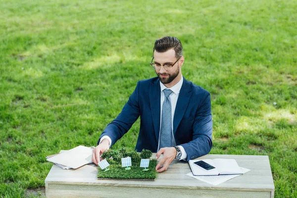 Uomo d'affari mettendo layout con batterie solari sul tavolo con notebook e smartphone mentre seduto in sedia ufficio nel parco — Foto stock