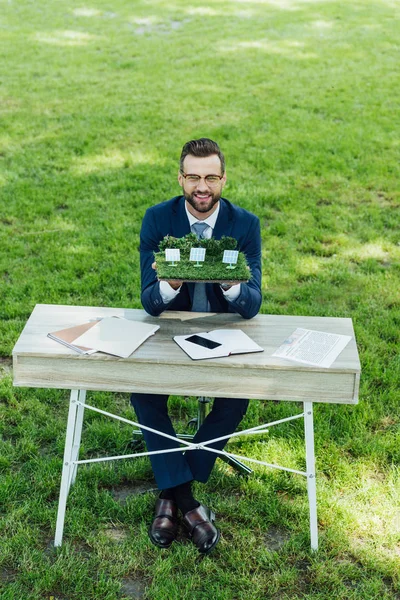 Visão de comprimento total do empresário segurando layout com baterias de sol na mesa com notebooks e smartphones enquanto sentado na cadeira de escritório no parque — Fotografia de Stock