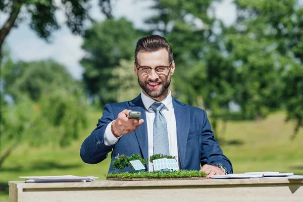 Young businessman with glasses sitting at table with sun batteries layout, looking at camera, smiling and presenting control panel — Stock Photo