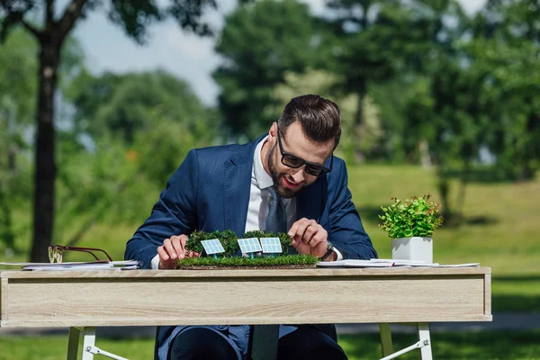 Joven hombre de negocios en gafas de sol sentado a la mesa con el diseño de baterías de sol y maceta con planta - foto de stock