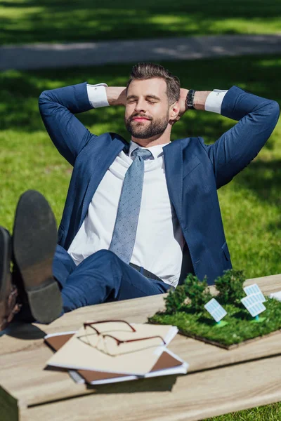 Молодий бізнесмен розслабляється, сидячи за столом з сонячними батареями планування, окуляри і блокноти, схрещені лаги і покласти руки за голову — стокове фото