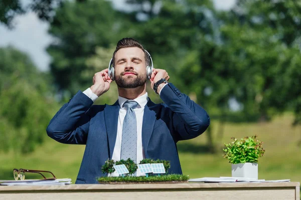 Joven hombre de negocios sentado en la mesa con el diseño de baterías de sol y maceta, relajante mientras escucha música con auriculares - foto de stock