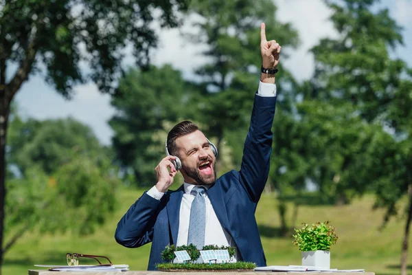 Возбужденный молодой бизнесмен сидит за столом с солнечными батареями макет и цветочный горшок, слушая музыку с наушниками и положить руку в воздух — стоковое фото