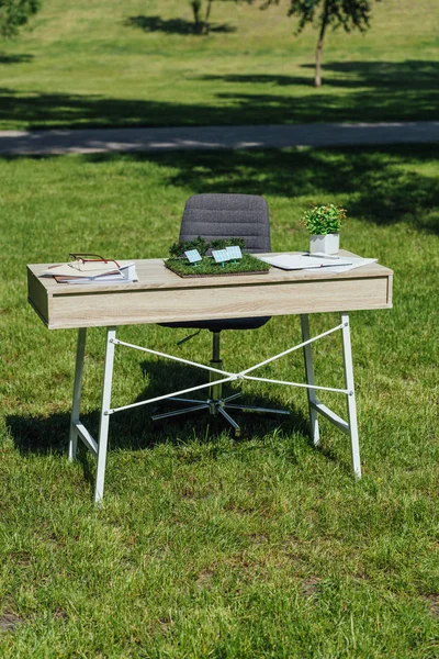 Tisch mit Sonnenbatterien, Flowepot, Notebooks und Brille in der Nähe eines schwarzen Bürostuhls im Park — Stockfoto