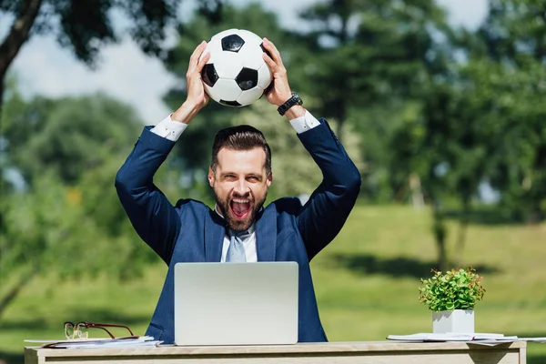 Jovem empresário sentado à mesa com laptop, vaso e óculos, sorrindo e segurando futebol sobre a cabeça — Fotografia de Stock
