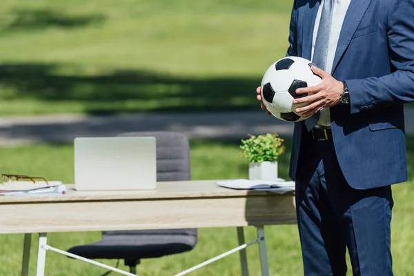 Cropped view of businessman holding soccer ball while standing in park near table with laptop and flowerpot — Stock Photo