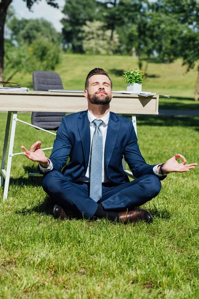 Бизнесмен сидит и медитирует на траве возле стола в парке — стоковое фото