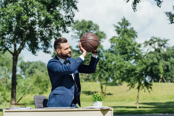 Молодой бизнесмен, занимающийся баскетболом, стоя в парке рядом со столом с цветочным горшком — стоковое фото