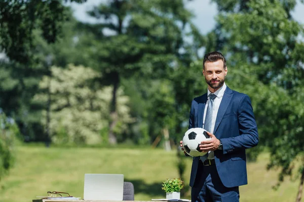 Young businessman holding football while standing in park near table with laptop and flowerpot and looking at camera — Stock Photo