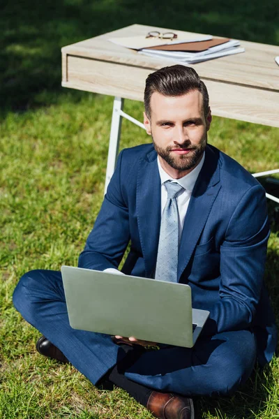 Young businessman sitting on grass in park with laptop near table and looking at camera — Stock Photo