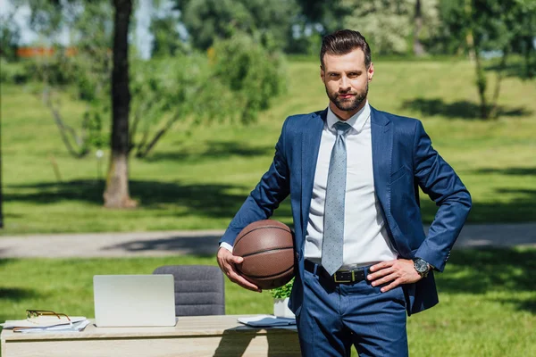 Homem de negócios em pé mesa limpa no parque, segurando basquete e olhando para a câmera — Fotografia de Stock
