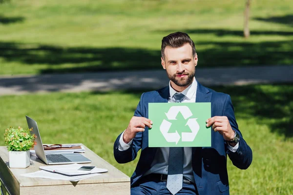 Young businessman sitting in park near table with laptop, holding card with recycle sign and looking at camera — Stock Photo