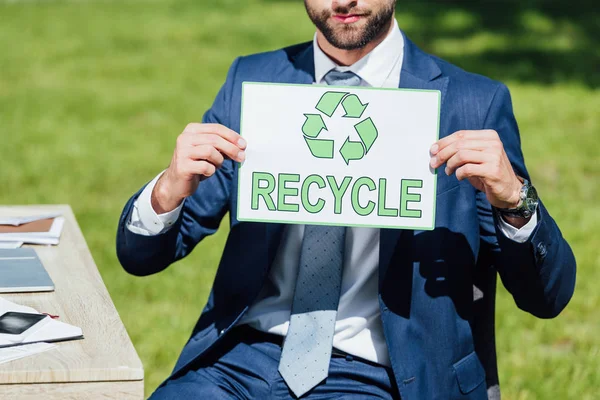 Vue recadrée de l'homme d'affaires assis près de la table et montrant la carte avec panneau de recyclage — Photo de stock