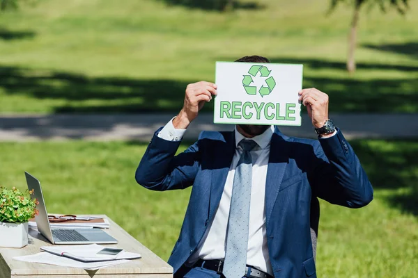 Young businessman sitting near table and covering face with card with recycle lettering — Stock Photo