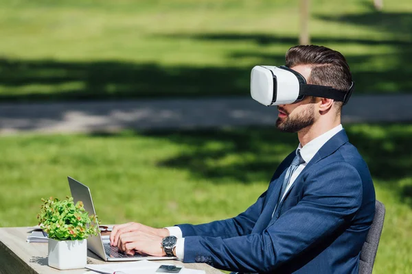 Joven hombre de negocios usando el ordenador portátil mientras está sentado en los auriculares VR en el parque - foto de stock