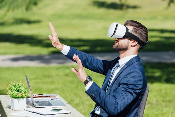 Jovem empresário em VR headset gesticulando enquanto se senta mesa com laptop, cadernos e vaso no parque — Fotografia de Stock