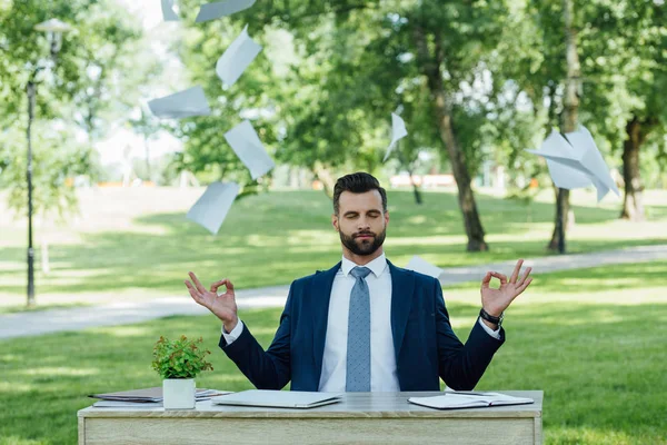 Молодой бизнесмен, сидящий в парке за столом и медитирующий возле летающих бумаг — стоковое фото