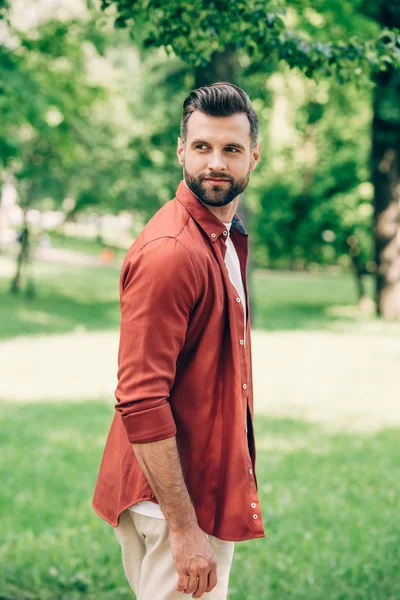 Joven hombre guapo en camisa roja mirando hacia otro lado mientras está de pie en el parque - foto de stock