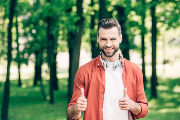 Jeune homme en chemise rouge debout dans le parc avec des écouteurs sur le cou et montrant les pouces vers le haut — Photo de stock