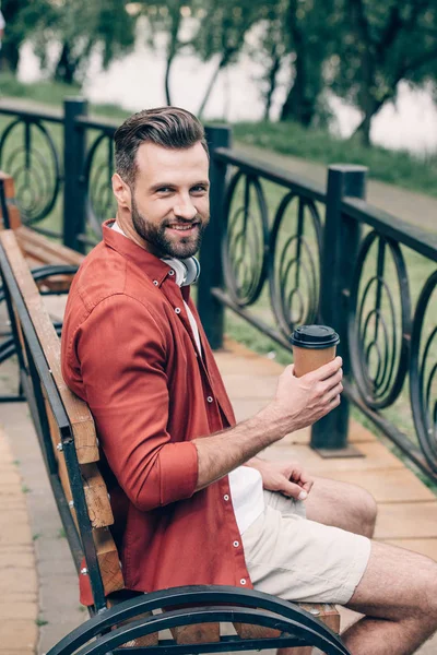 Jovem de camisa vermelha sentado no banco, segurando café para ir, sorrindo e olhando para a câmera — Fotografia de Stock