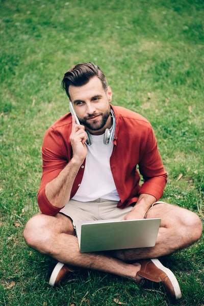 Joven sentado en la hierba en el parque, hablando en el teléfono inteligente y utilizando el ordenador portátil - foto de stock