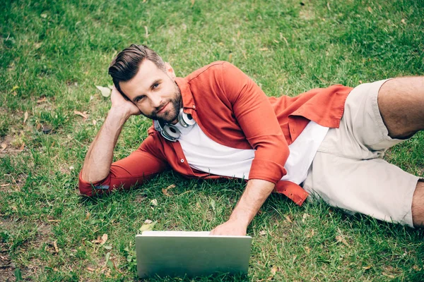 Молодой человек лежит на траве в парке, использует ноутбук и смотрит в камеру — стоковое фото
