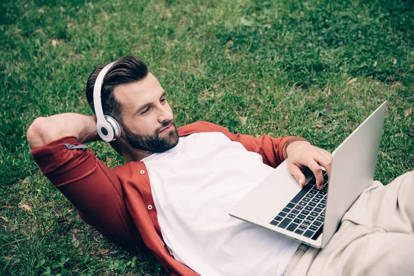 Молодой человек лежит на траве в парке, используя ноутбук и слушая музыку на наушниках — стоковое фото