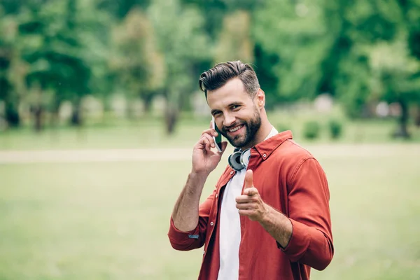 Young man standing in park, talking on smartphone, smiling, looking and pointing with finger at camera — Stock Photo