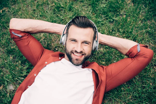Jeune homme allongé sur l'herbe avec les mains derrière la tête, écoutant de la musique, souriant et regardant la caméra — Photo de stock