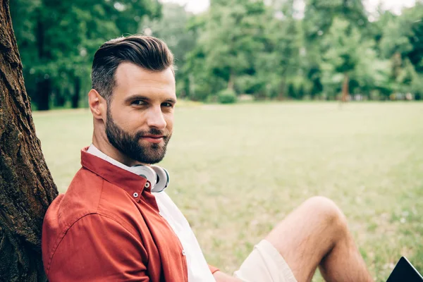 Young man sitting in park near tree and looking at camera — Stock Photo