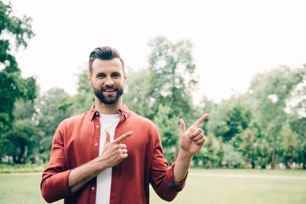 Mann in rotem Hemd steht im Park, blickt in Kamera und zeigt mit den Fingern — Stockfoto