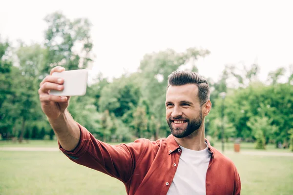 Bell'uomo in piedi nel parco, sorridente e prendendo selfie — Foto stock