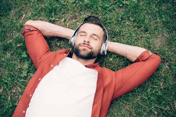 Joven con los ojos cerrados tumbado en la hierba con las manos detrás de la cabeza y escuchando música - foto de stock