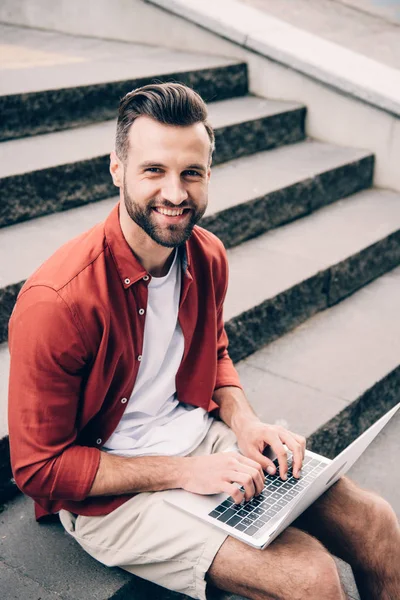 Young man sitting on stone stairs, using laptop, smiling and looking at camera — Stock Photo