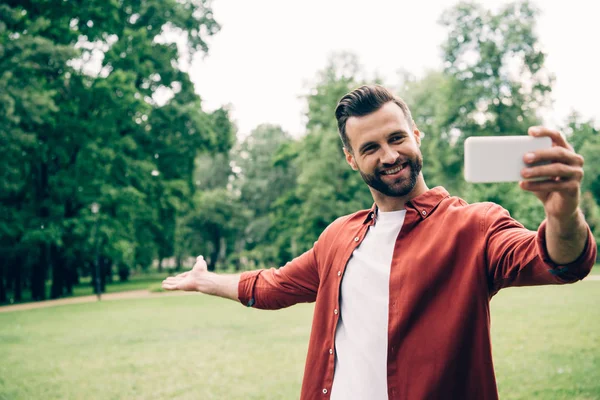 Красивый мужчина, стоящий в парке, улыбающийся, делающий селфи и жестикулирующий рукой — стоковое фото