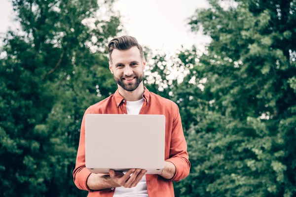 Junger Mann steht im Park, hält Laptop in der Hand und blickt in Kamera — Stockfoto