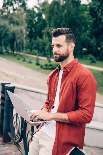 Jeune homme utilisant un ordinateur portable près du parc et regardant loin — Photo de stock