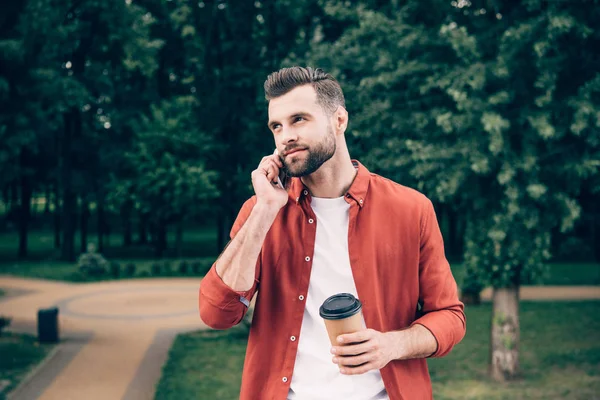 Молодий чоловік розмовляє по смартфону і тримає каву, щоб піти, стоячи в парку — стокове фото