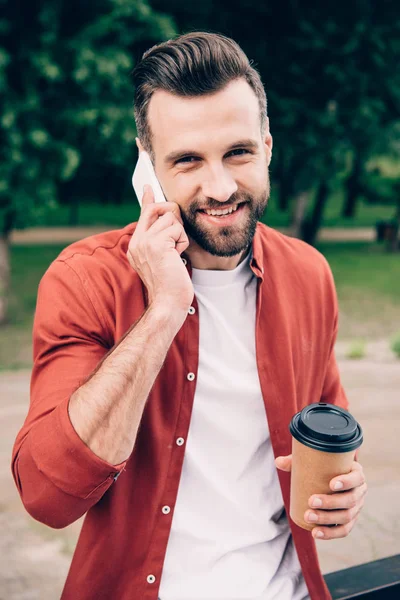 Hombre guapo hablando en el teléfono inteligente, sosteniendo el café para ir y mirando a la cámara mientras está de pie en el parque - foto de stock