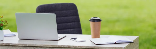 Panoramic shot of table with laptop, smartphone, notebook and coffee to go near office chair in park — Stock Photo