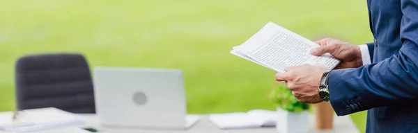 Panoramic shot of man holding newspaper in park near white table with office stuff — Stock Photo