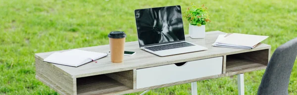 Plano panorámico de mesa blanca con portátil, smartphone, café para llevar, cuadernos y planta en parque - foto de stock
