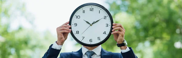 Panoramic shot of businessman covering face with clock while standing in park — Stock Photo