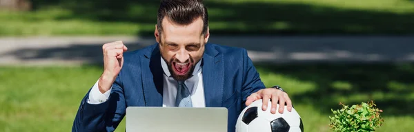 Panoramic shot of young businessman watching on laptop, cheering on team and holding soccer ball — Stock Photo