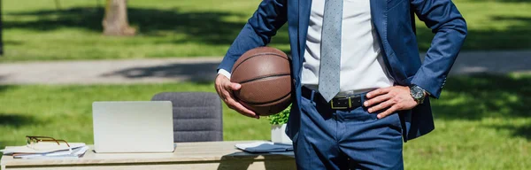 Panoramic shot of businessman holding basketball while standing in park near white office table with laptop — Stock Photo