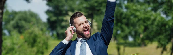 Panoramic shot of excited young businessman listening to music and rising hand in air — Stock Photo