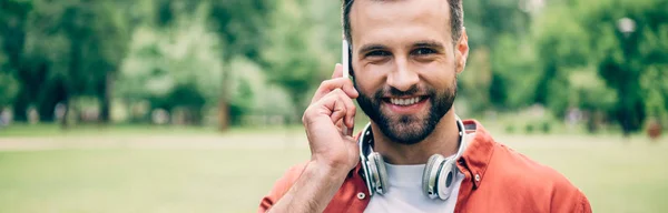 Панорамный снимок улыбающегося молодого человека, разговаривающего на смартфоне и смотрящего в камеру — стоковое фото