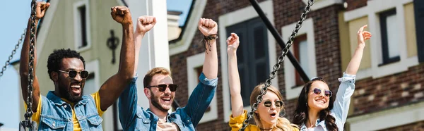 Panoramic shot of cheerful group of multicultural friends in sunglasses celebrating triumph — Stock Photo