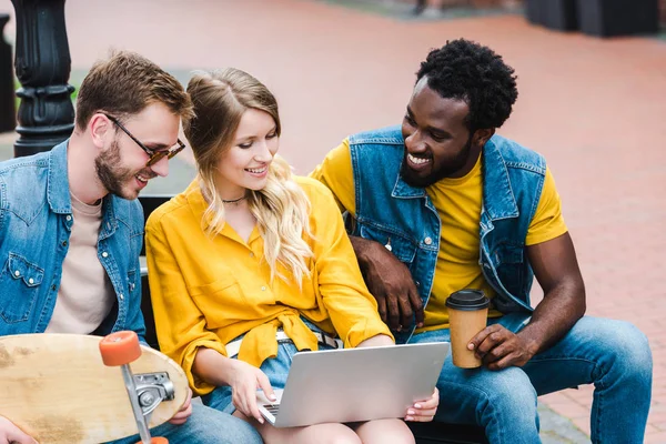 Cheerful young woman using laptop near happy multicultural men — Stock Photo