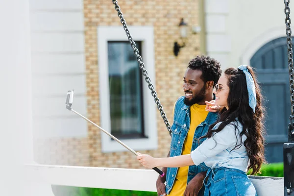 Attrayant femme montrant signe de paix près beau homme afro-américain tout en prenant selfie et tenant selfie bâton — Photo de stock